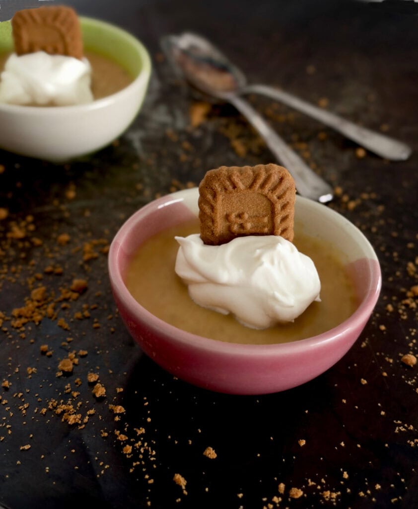 Pumpkin spice pots de crème in a pink ramekin, topped with whipped cream and a Biscoff cookie, with cookie crumbs and vintage spoons on a dark tray.
