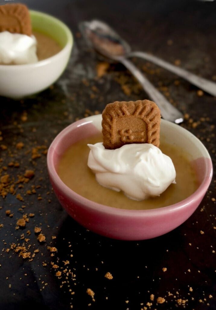 Pumpkin spice pots de crème in a pink ramekin, topped with whipped cream and a Biscoff cookie, with cookie crumbs and vintage spoons on a dark tray.